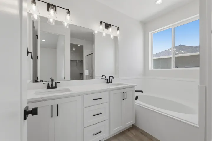Double vanity with integrated sinks and black fixtures, white cabinetry, large mirror, drop-in bathtub, and a large window