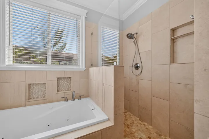 Tiled shower with handheld shower head and built-in niche, white soaking tub with integrated jets, and dual windows