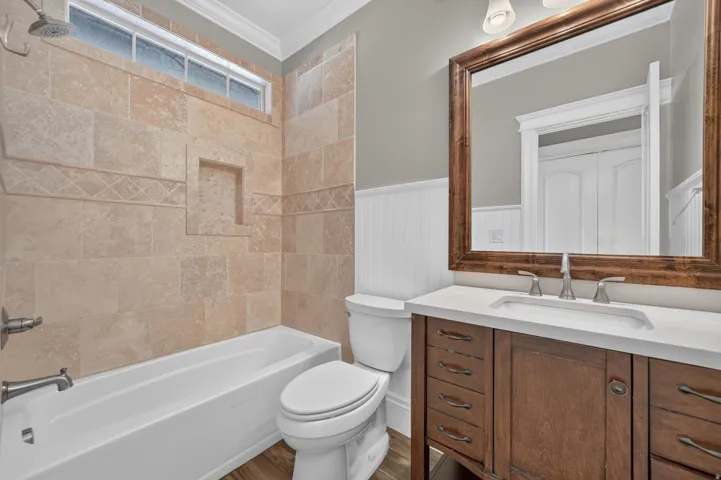 Wood-finish flooring, white vanity with integrated sink, white countertop, dark wood-framed mirror, and tiled shower surround with accent tile and niche