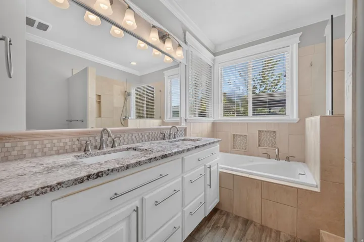 Dual vanity with stone countertop, large mirror with vanity lighting, integrated bathtub, tiled shower, and window with blinds