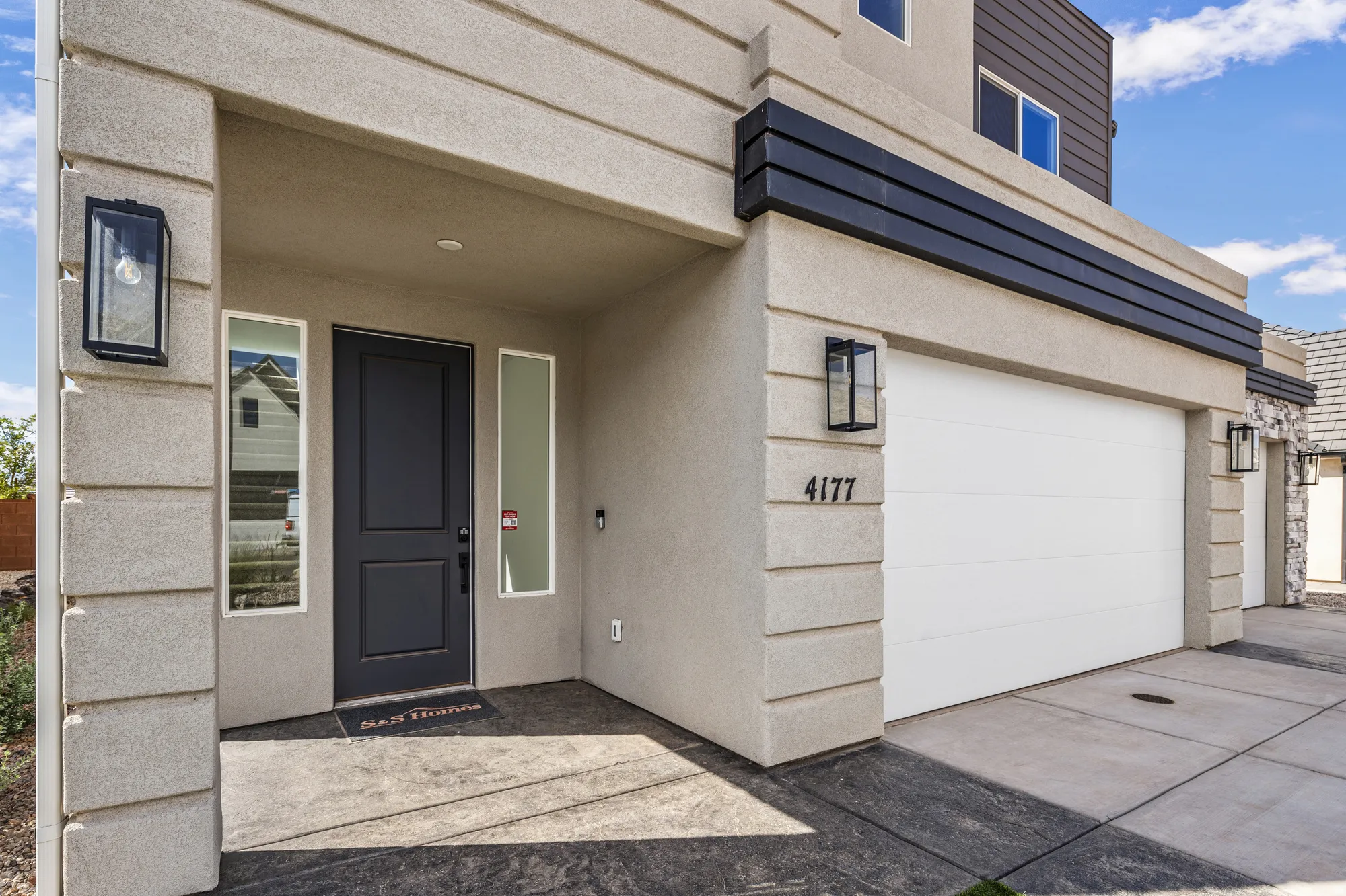 Cream stucco exterior with decorative horizontal banding, dark gray entry door, two vertical sidelight windows, and a white garage door with house numbers