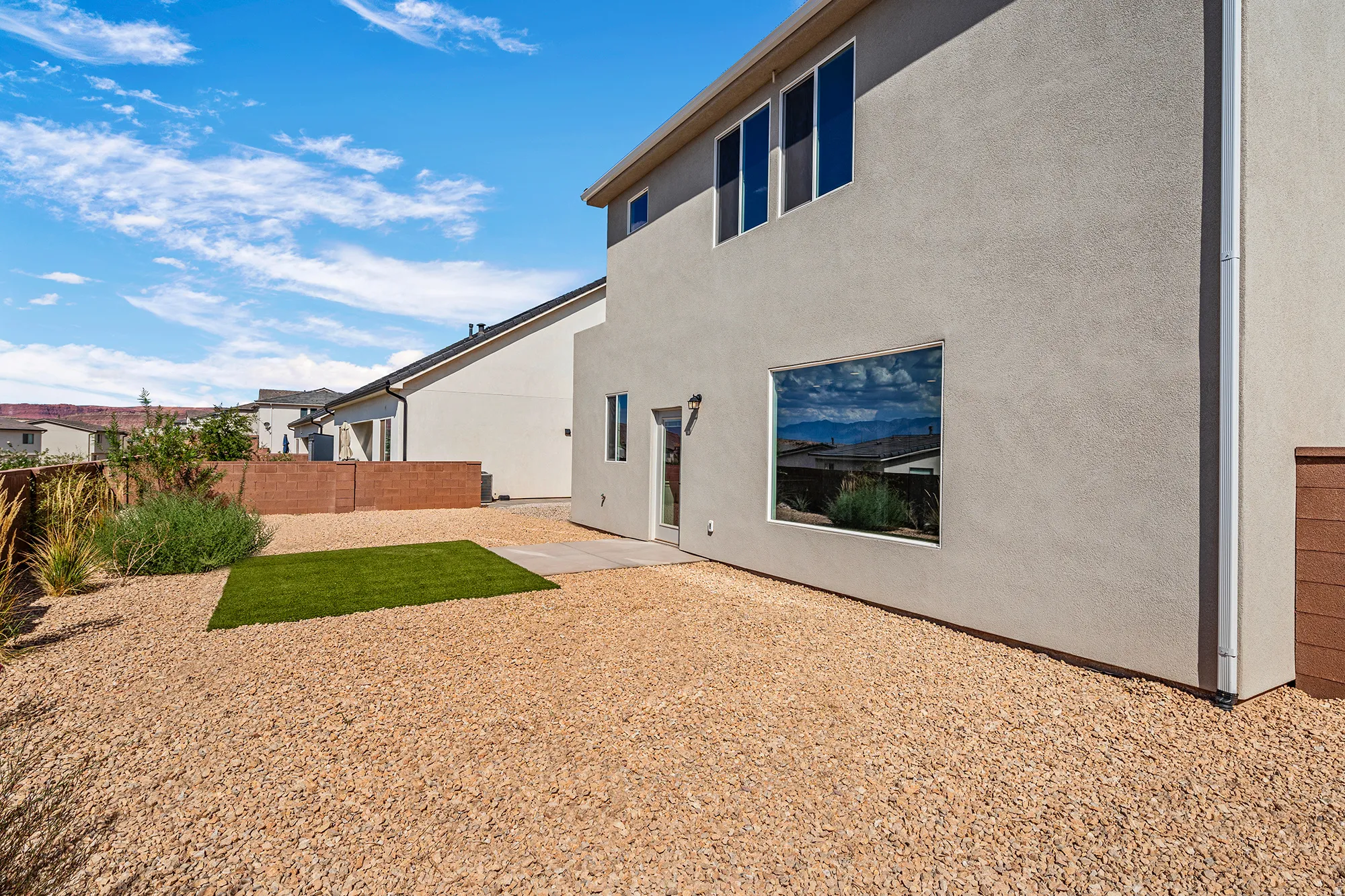 Stucco exterior with multiple windows, featuring a gravel yard with a synthetic turf area, and a low brick retaining wall