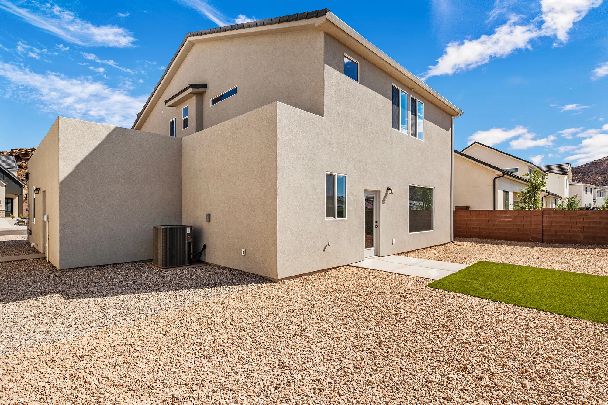 Stucco exterior with multiple windows, leading to an expansive gravel yard featuring an artificial turf area and a concrete patio