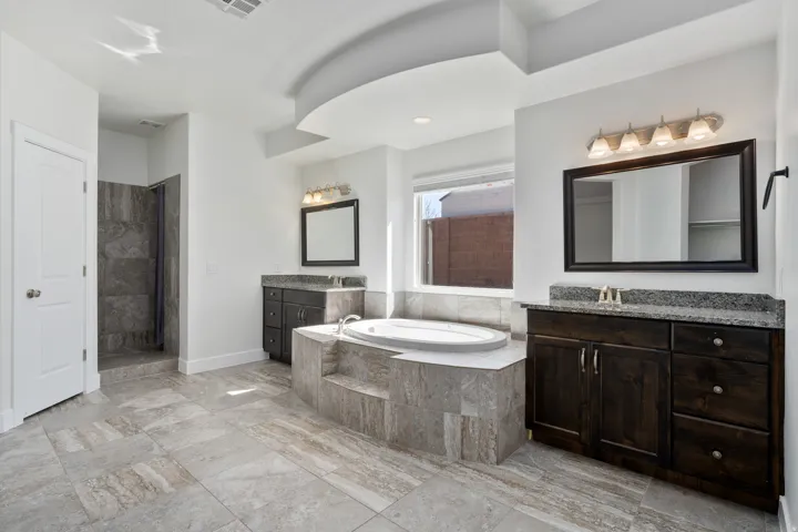 Spacious bathroom featuring an oval soaking tub, dual dark wood-finish vanities with stone countertops, a walk-in shower with tiled walls, and large format floor tiling