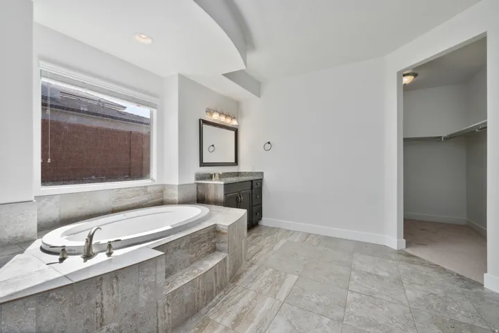 Built-in soaking tub with tile surround, single vanity with dark cabinetry and stone countertop, framed mirror, and recessed lighting