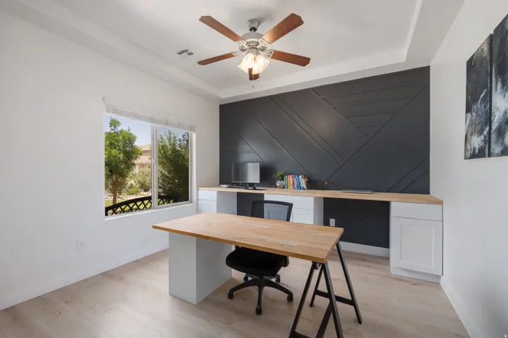 Recessed ceiling with a central fan fixture, wood-finish flooring, a large window, a dark accent wall with geometric paneling, and built-in desk cabinetry with a light wood-finish countertop