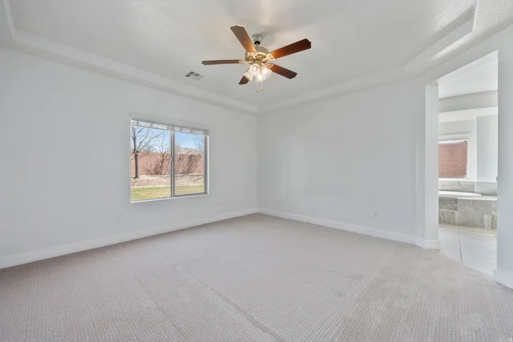 Tray ceiling, ceiling fan with light fixture, window with blinds, light-toned carpet, and an adjacent tiled bathroom area with a built-in tub