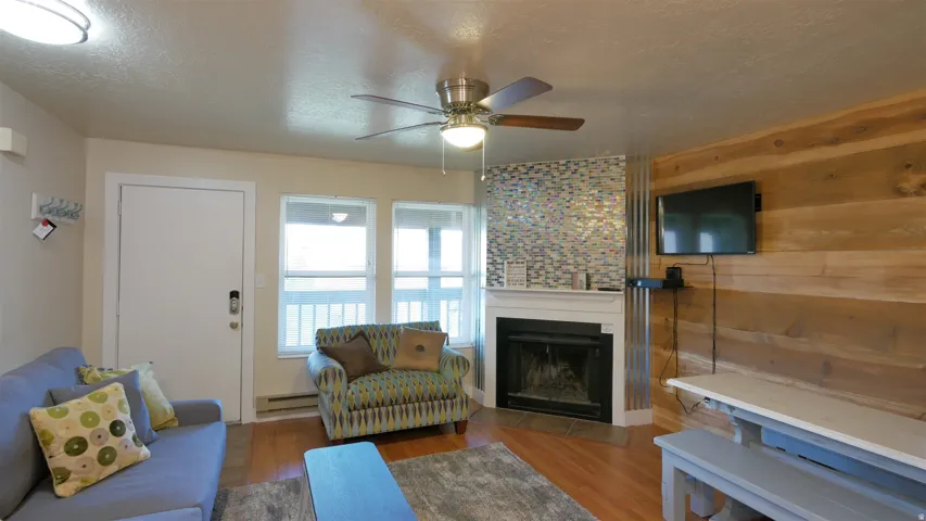 Wood-finish flooring, built-in fireplace with tile surround, wood-paneled accent wall, ceiling fan with integrated lighting, and multiple windows