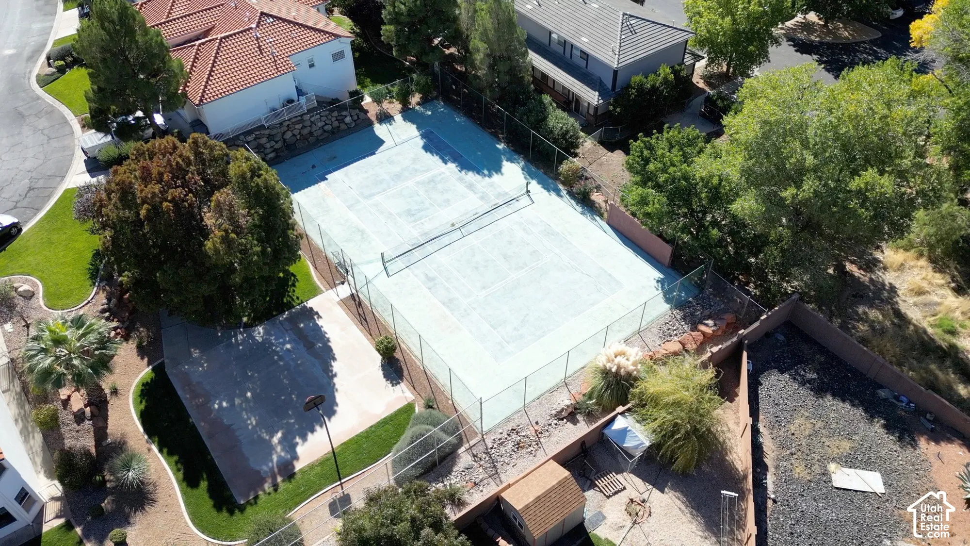 Full-size tennis court with perimeter fencing, surrounded by mature trees and paved pathways