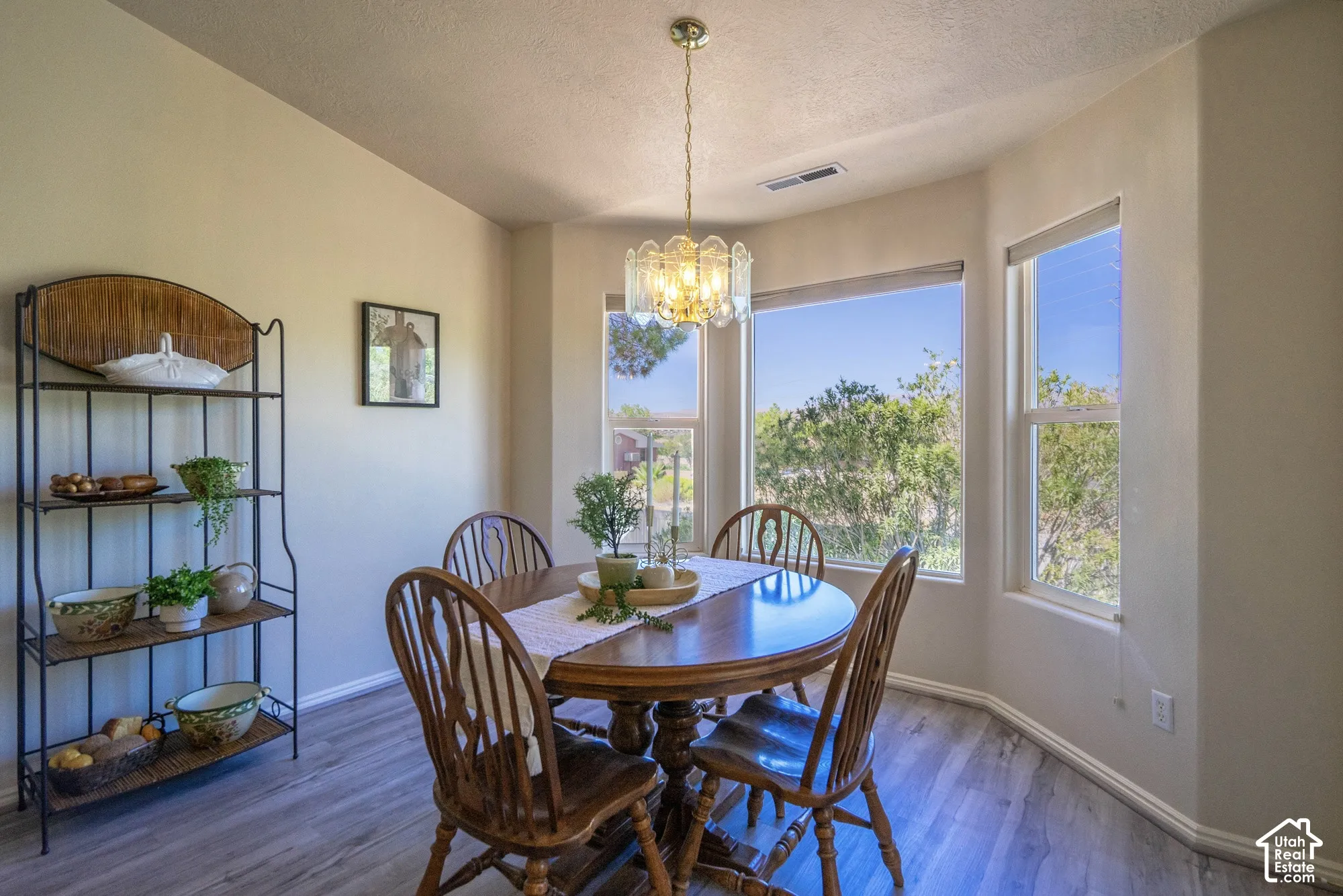 Bay windows with roller shades, a brass chandelier, and wood-finish flooring