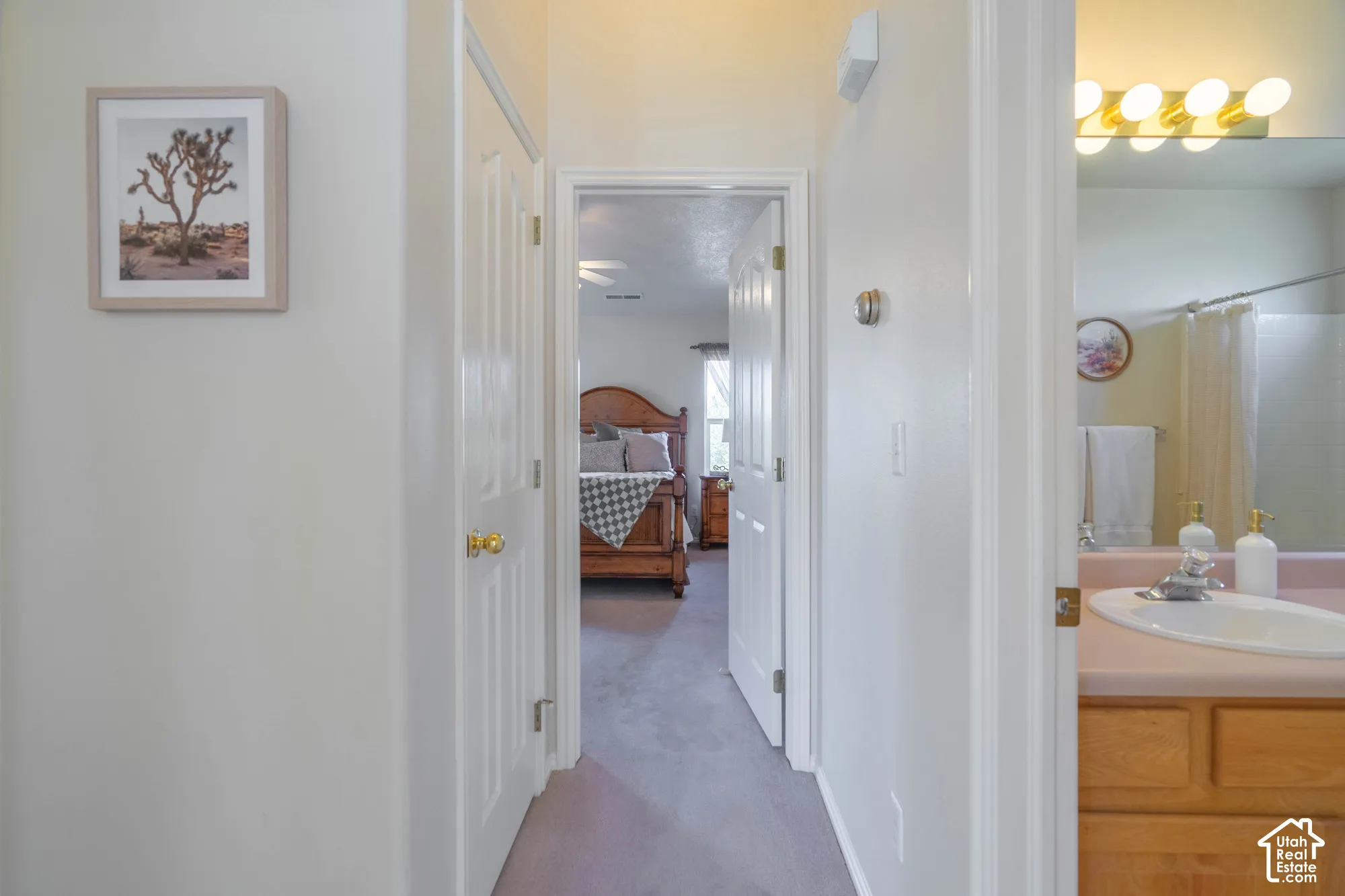 Light-toned interior walls, wood-finish vanity with countertop and integrated sink, full-width mirror with overhead lighting, and an additional doorway revealing a bedroom with a ceiling fan
