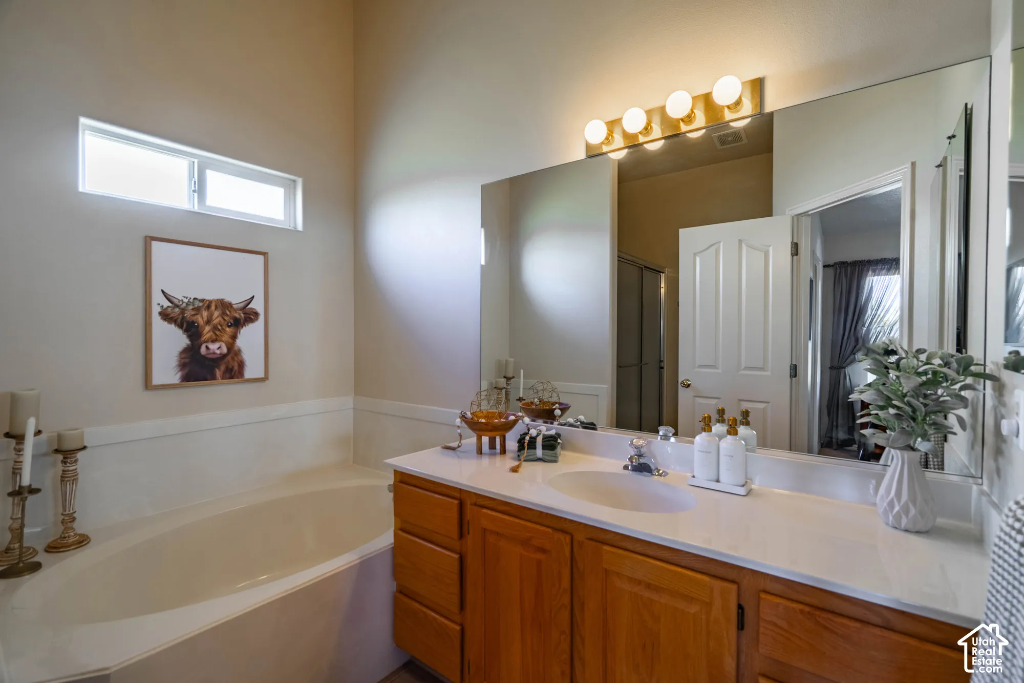 Soaking tub with a high transom window, wood-finish vanity with an integrated sink, and an expansive wall-mounted mirror with overhead lighting