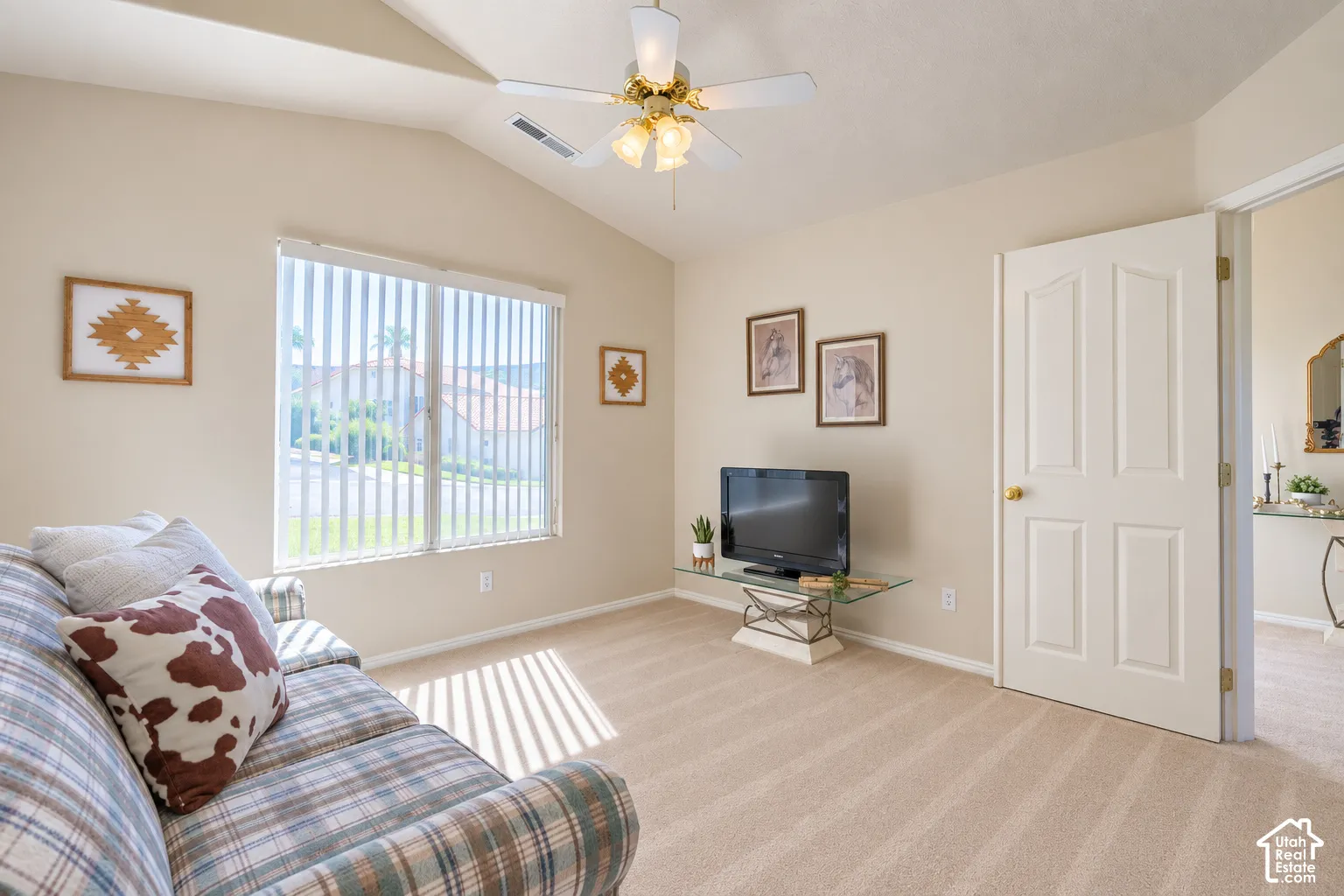 Vaulted ceiling, ceiling fan with light fixture, large window with vertical blinds, neutral tone walls, and plush carpet flooring