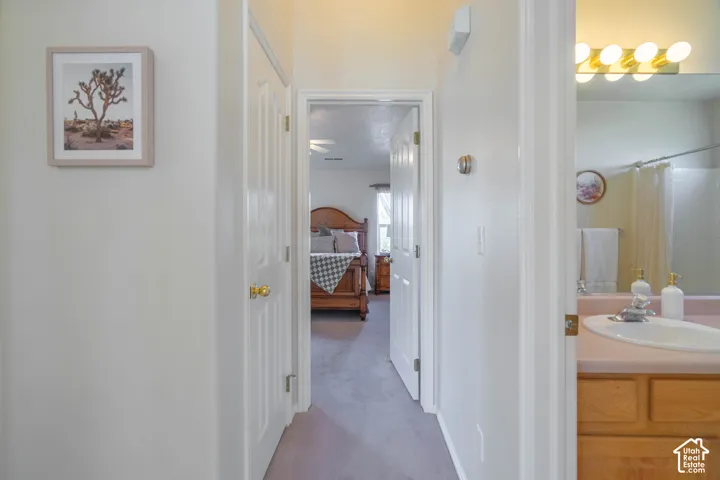 Light-toned interior walls, wood-finish vanity with countertop and integrated sink, full-width mirror with overhead lighting, and an additional doorway revealing a bedroom with a ceiling fan
