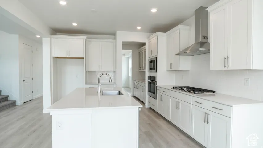 Kitchen featuring white cabinetry, light-toned countertops, a stainless steel range hood, built-in double ovens, and a central island with an integrated sink