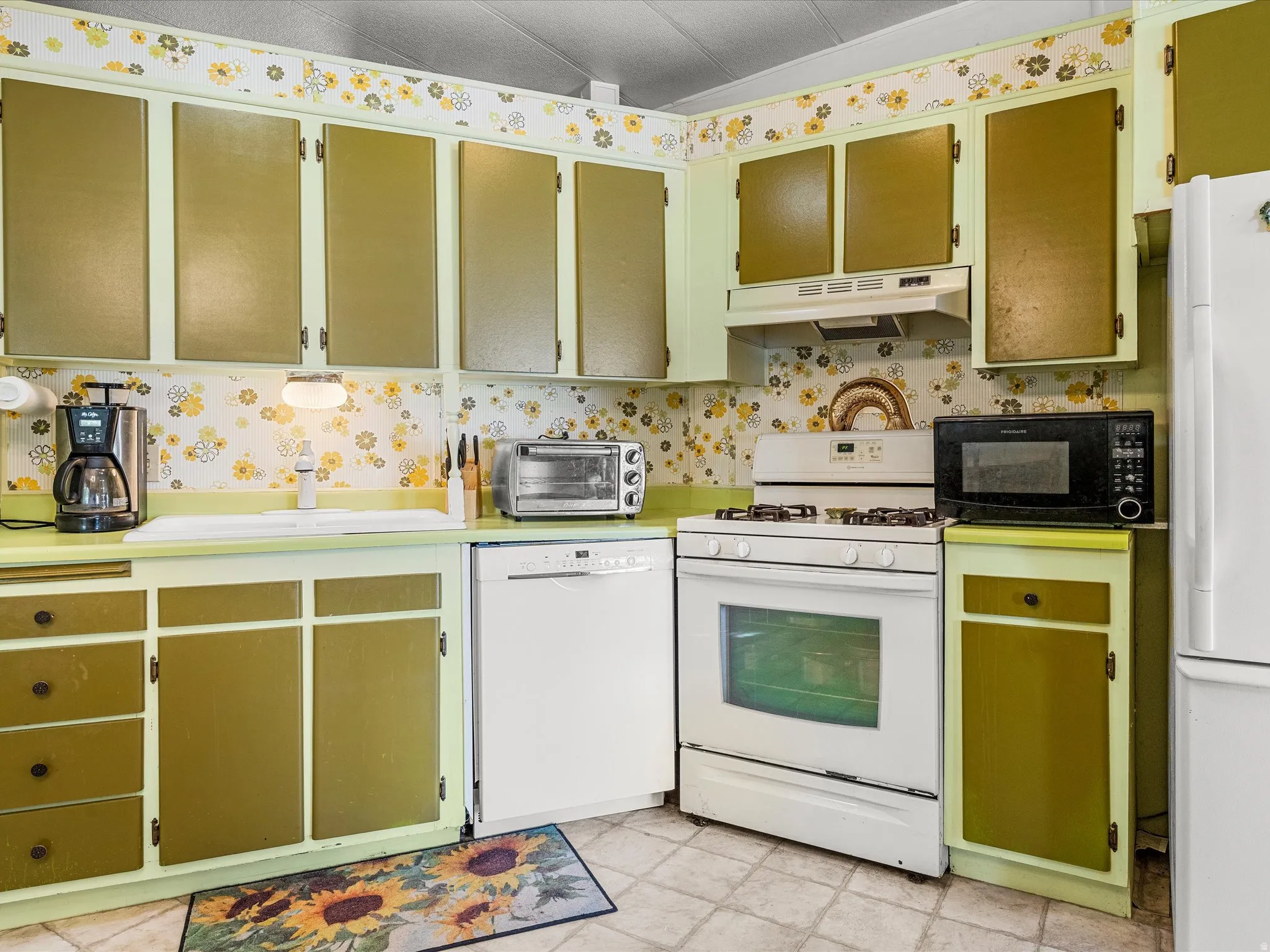 Kitchen featuring white appliances, upper and lower cabinetry, a white sink, and patterned wallpaper