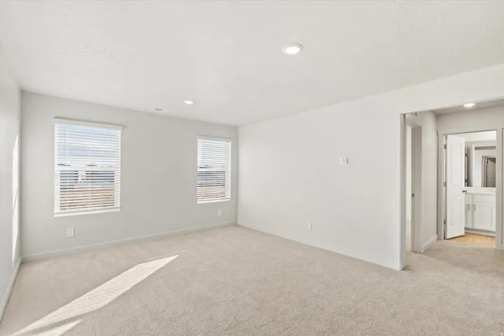 Spacious room with plush carpeting, two windows with blinds, recessed lighting, and a white interior door leading to an adjacent bathroom with a vanity