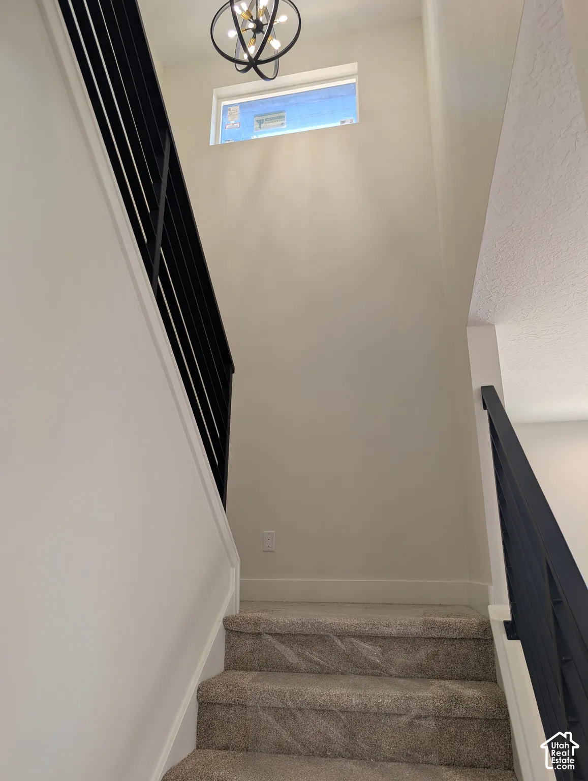 Carpeted staircase with black vertical baluster railing, featuring a large modern chandelier and a transom window