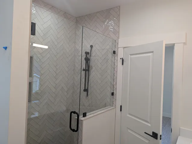 Glass-enclosed shower with herringbone-patterned tile, matte black shower fixtures, and a coordinating matte black door handle
