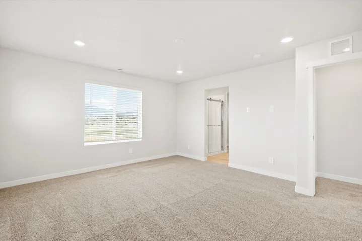 Bright, open bedroom featuring neutral carpeting, recessed ceiling lighting, and a large window with plantation shutters allowing natural light throughout the space