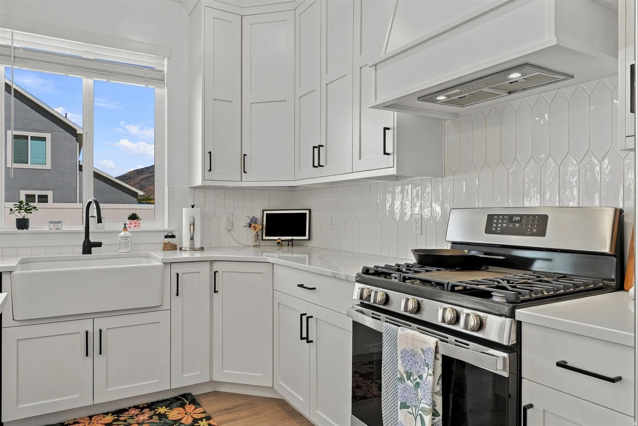 White shaker cabinetry, stone countertops, farmhouse sink with gooseneck faucet, stainless steel gas range, and a decorative tile backsplash