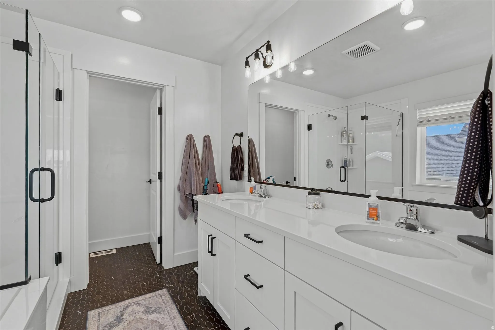 Dual vanity with white cabinetry and integrated sinks, a large wall-mounted mirror, a glass-enclosed shower, and hexagonal tile flooring