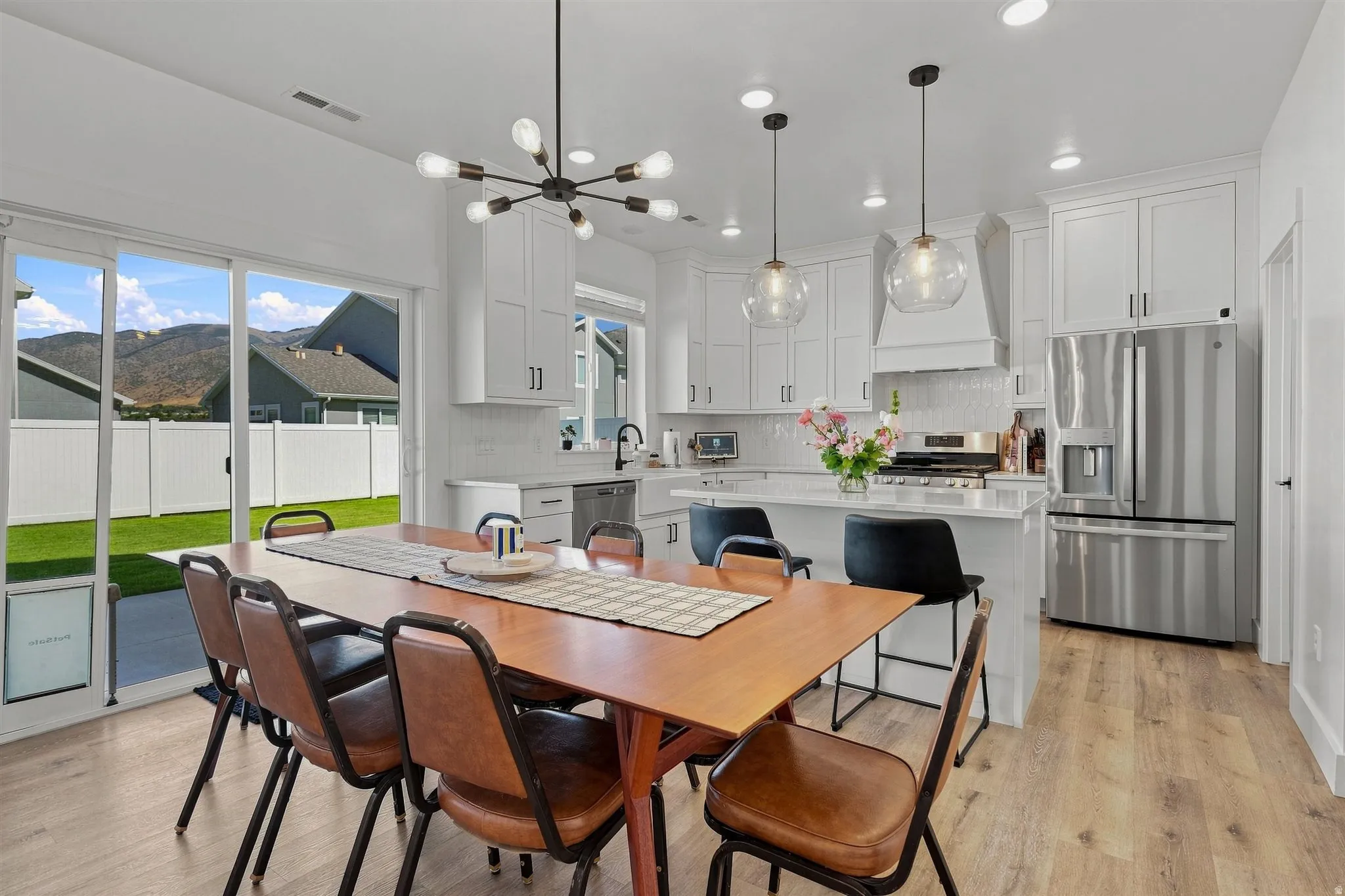 Bright open-concept kitchen and dining area featuring a modern chandelier, wood-finish flooring, white cabinetry, and stainless steel appliances