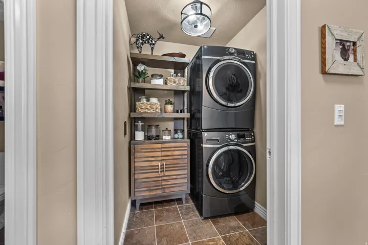 Laundry area featuring stacked front-load machines, a built-in wood-finish shelving unit, a wood-finish cabinet with horizontal detailing, and tile flooring