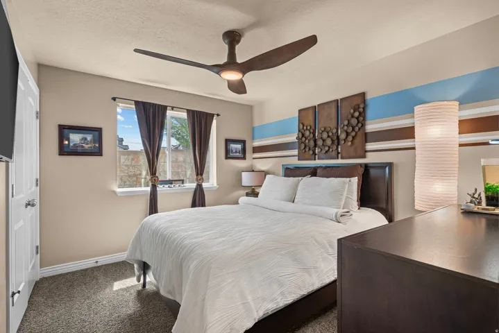 Ceiling fan with integrated lighting, window with dark brown draperies, textured wall art, dark wood-finish dresser, and carpet flooring