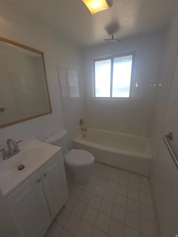 White subway tile shower surround, white vanity with integrated sink, white tub, tile flooring, and a window