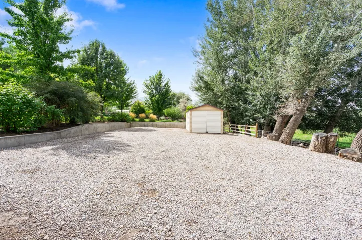 Expansive gravel area with mature trees, a detached storage shed, and a concrete retaining wall