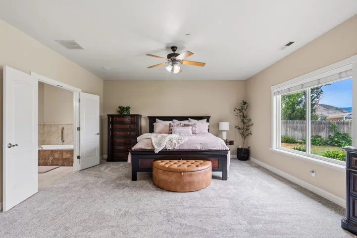 Carpeted flooring throughout, double-door entry to a tiled bathroom with a built-in tub, a large window, and a ceiling fan