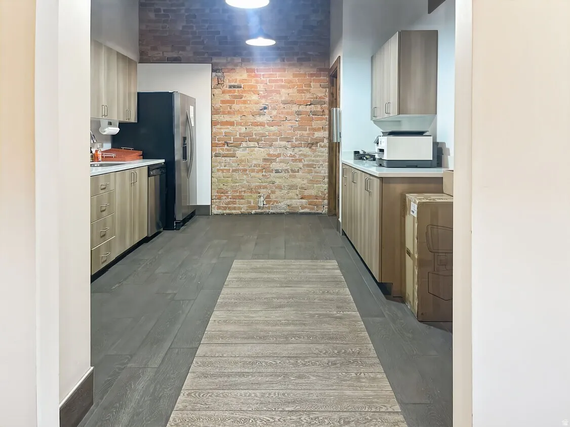 Exposed brick wall, wood-finish flooring, light-toned cabinetry, stainless steel refrigerator, and recessed lighting