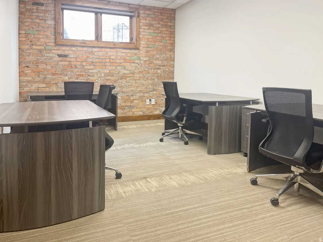 Exposed brick wall, wood-frame window, light-toned patterned carpeting, white painted walls, and wood-finish office desks