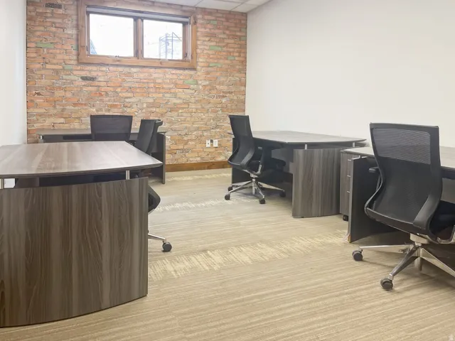 Exposed brick wall, wood-frame window, light-toned patterned carpeting, white painted walls, and wood-finish office desks