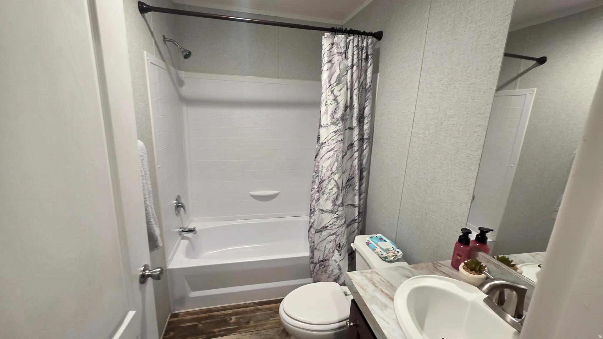 White bathtub with an integrated shower, chrome fixtures, a wood-finish flooring, and a vanity with a drop-in sink