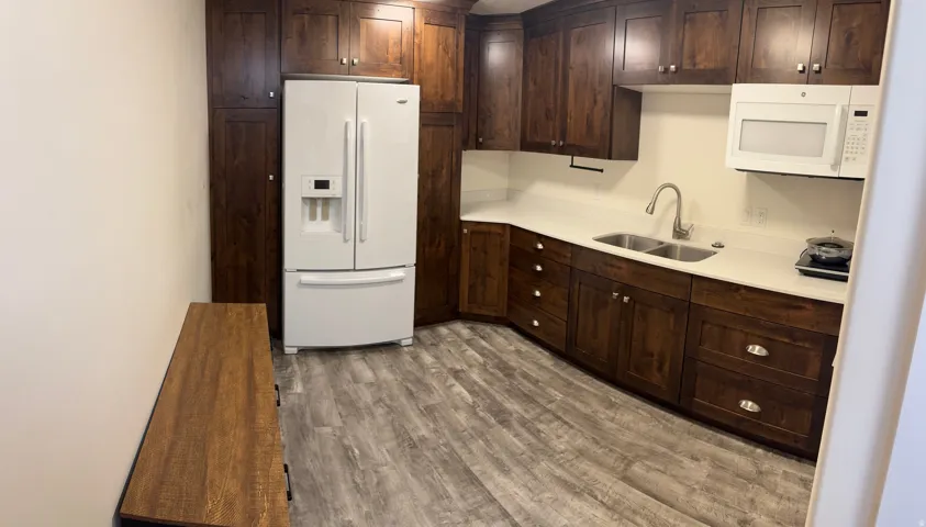Dark wood-finish cabinetry, white countertop, stainless steel double basin sink with gooseneck faucet, white microwave, and Vinyl flooring