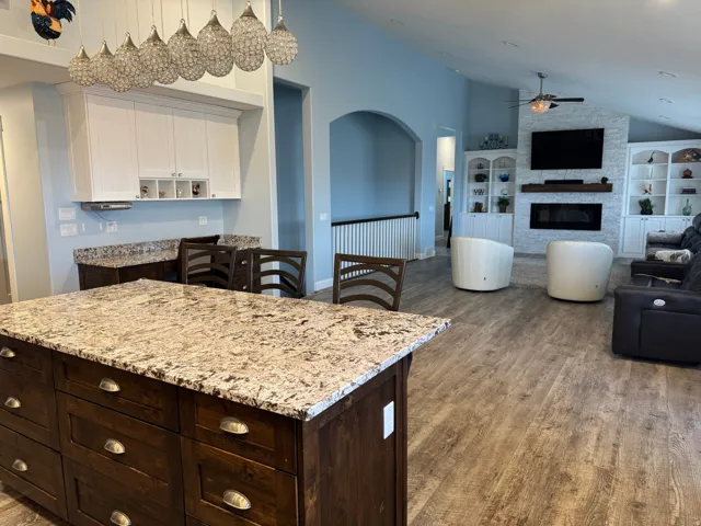 Open-concept kitchen and living area featuring a large granite-topped island with multiple drawers, white cabinetry with stainless steel appliances, Vinyl flooring throughout, and a fireplace with built-in shelving and media niche