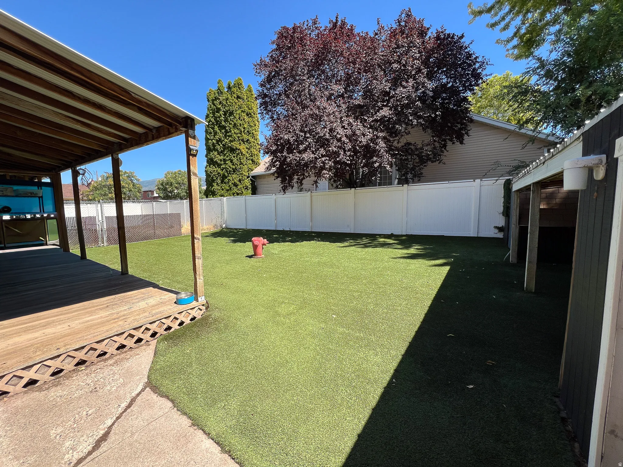 Expansive backyard featuring a covered deck, artificial turf, a white privacy fence, and mature trees