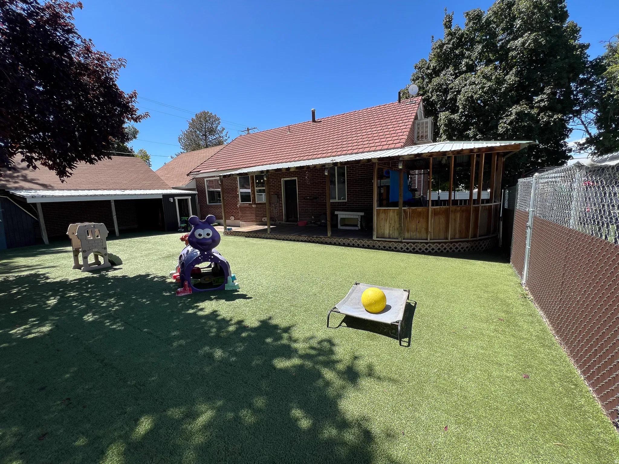 Expansive artificial turf backyard featuring a covered patio, brick exterior, and a chain link fence with privacy slats