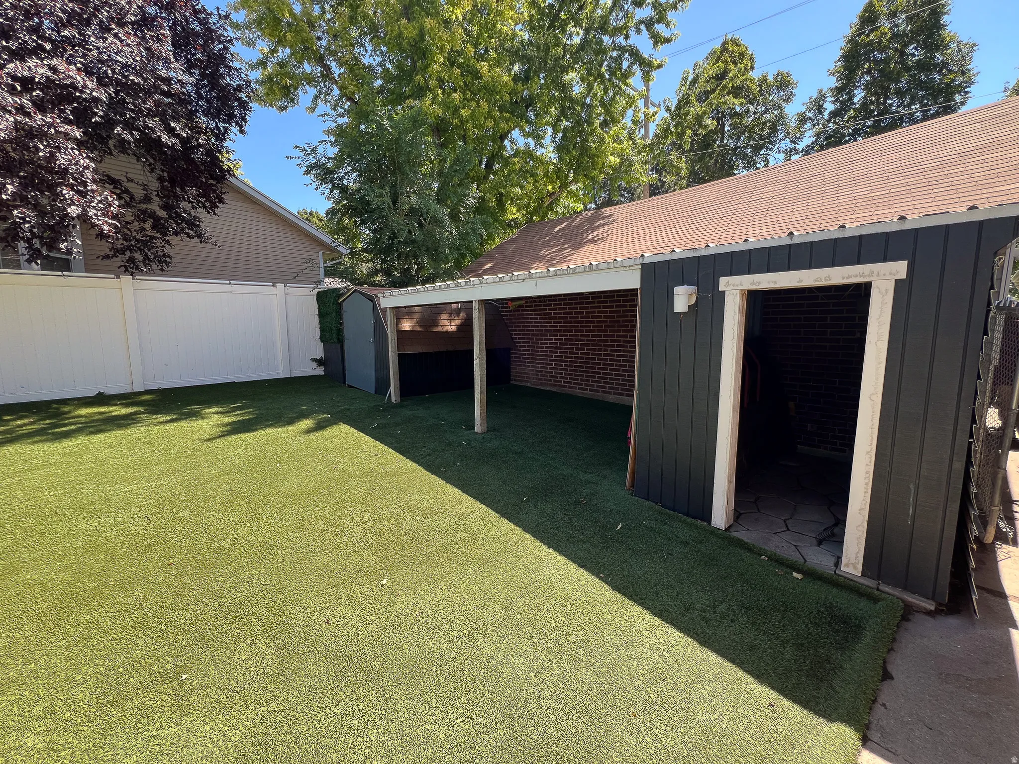 Expansive outdoor space featuring artificial turf, a white privacy fence, a detached shed, a covered patio area with brick siding, and a separate storage structure with dark paneling