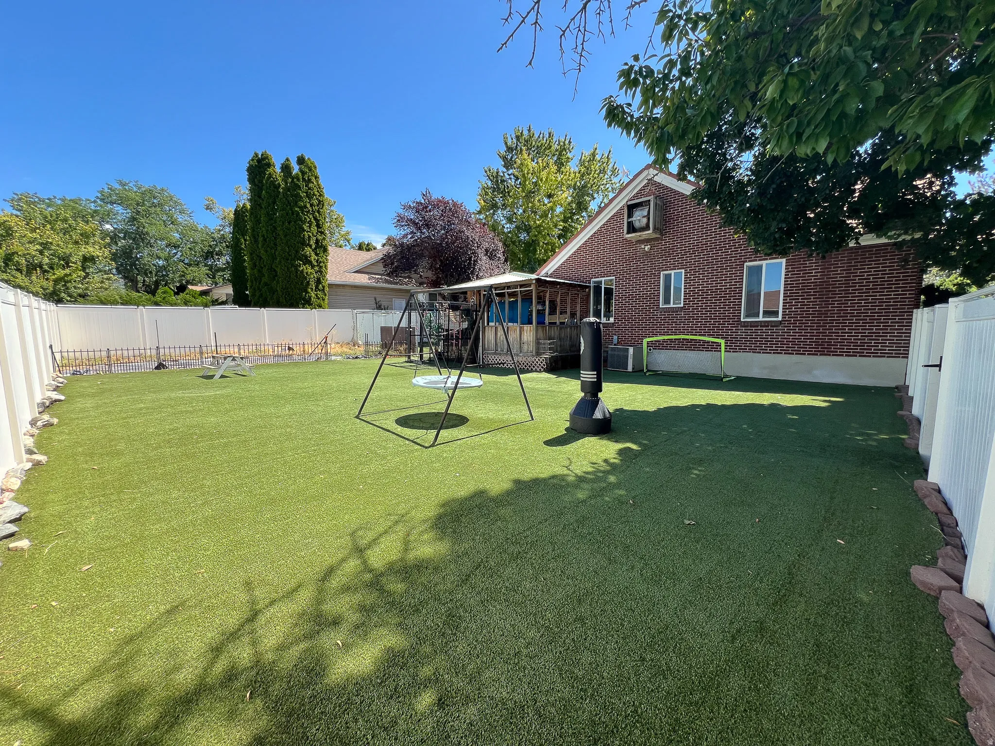 Expansive backyard featuring artificial turf, a brick exterior, white privacy fencing, a covered patio, and mature trees