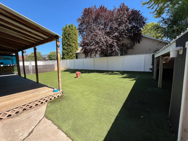 Expansive backyard featuring a covered deck, artificial turf, a white privacy fence, and mature trees