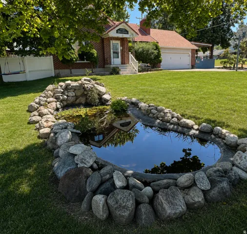 Manicured lawn with a stone-edged pond, brick exterior, white garage door, and a white fence