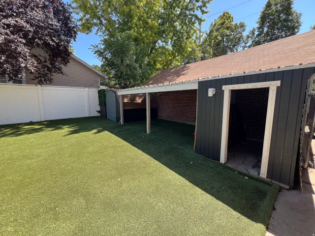 Expansive outdoor space featuring artificial turf, a white privacy fence, a detached shed, a covered patio area with brick siding, and a separate storage structure with dark paneling