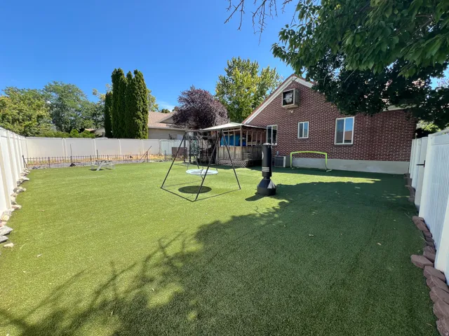 Expansive backyard featuring artificial turf, a brick exterior, white privacy fencing, a covered patio, and mature trees