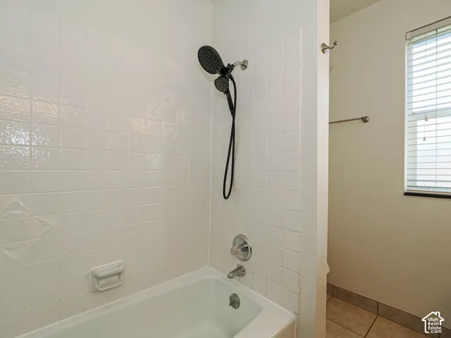 Bathtub enclosure featuring white wall-tile surround with patterned detailing, multi-function handheld shower head, integrated soap dish, and horizontal blind window treatment