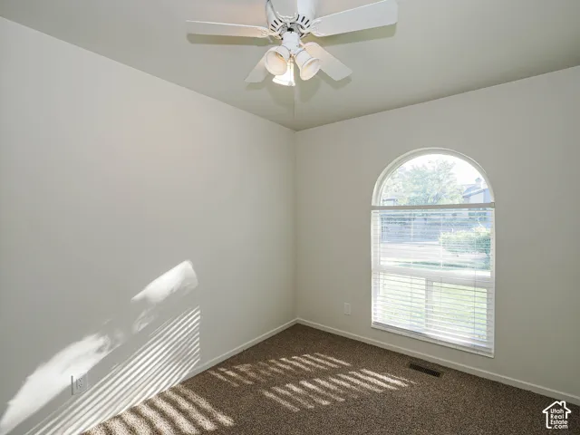 Neutral-toned room featuring a central ceiling fan with integrated lighting, textured carpeting, and a prominent arched window with horizontal blinds