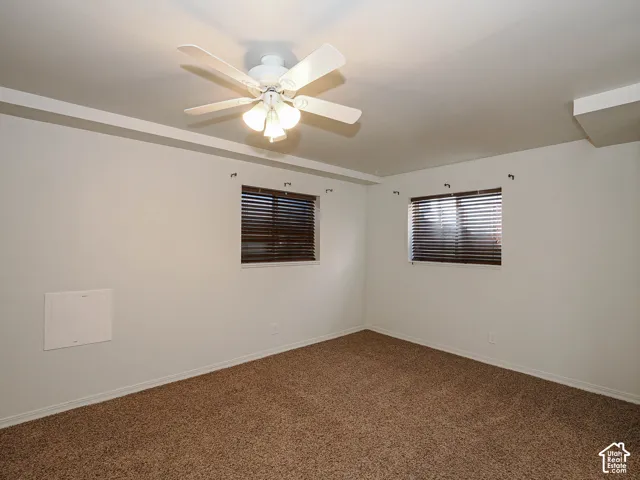 Neutral-toned interior space featuring textured carpeting, two windows with horizontal blinds, and a white ceiling fan with integrated lighting