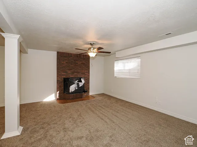 Brick fireplace featuring a metal insert, ceiling fan with integrated lighting, neutral carpeting, and a structural column